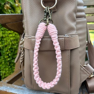 Może przedstawiać: Różowy pleciony brelok z pozłacanym metalowym zapięciem przymocowanym do brązowego skórzanego plecaka.