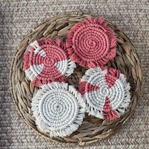 May include: Four round woven coasters with fringe. Two coasters are white with a pink center and two coasters are pink with a white center.