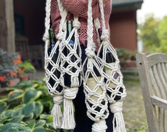 Puede incluir: Un porta botellas de vino de macramé blanco con cuatro botellas de vino en su interior. El porta botellas tiene borlas largas y se lleva sobre el hombro.