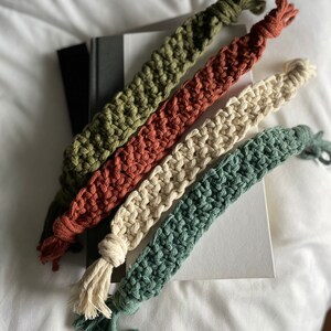 May include: Four crocheted bookmarks in different colors: green, red, cream, and dark green. The bookmarks are arranged on top of a closed book.
