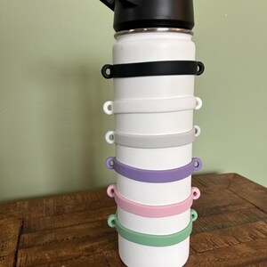 May include: A white water bottle with a black lid and a series of colorful silicone bands. The bands are black, white, gray, purple, pink, and green. The bottle is on a wooden surface.