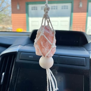 May include: A pink Himalayan salt crystal is suspended in a white macrame hanging, with a white wooden bead at the bottom. The hanging is attached to a rearview mirror in a car.