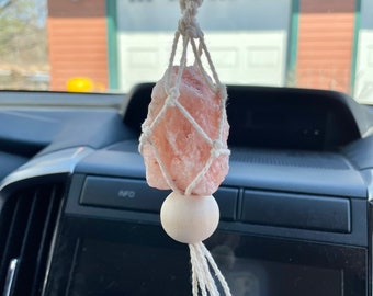 Puede incluir: Un cristal de sal rosa del Himalaya está suspendido en una macramé blanca colgante, con una cuenta de madera blanca en la parte inferior. La colgante está unida a un espejo retrovisor en un coche.