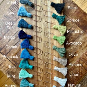 May include: A set of 14 gold paper clips with colorful tassels. The tassels are in shades of blue, green, and beige. The colors are labeled with names like "Steel", "Sky", "Deep Sea", "Indigo", "Cobalt", "Peacock", "Teal", "Mint", "Agave", "Black", "Spruce", "Pistachio", "Avocado", "Olive", "Almond", "Linen", "Natural", and "White".