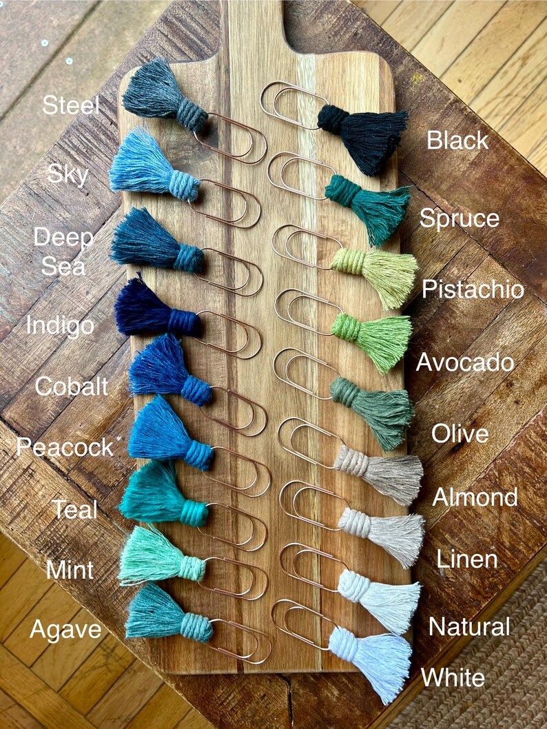 May include: A set of 14 gold paper clips with colorful tassels. The tassels are in shades of blue, green, and beige. The colors are labeled with names like "Steel", "Sky", "Deep Sea", "Indigo", "Cobalt", "Peacock", "Teal", "Mint", "Agave", "Black", "Spruce", "Pistachio", "Avocado", "Olive", "Almond", "Linen", "Natural", and "White".