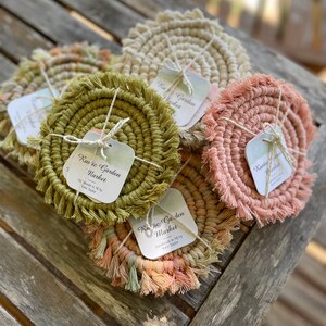 May include: Set of six handmade crocheted coasters with fringe. The coasters are in various colors, including green, pink, and beige. Each coaster has a small square tag with the text "Rustic Garden Market" and "Handmade in MI by Lazie Taylor".