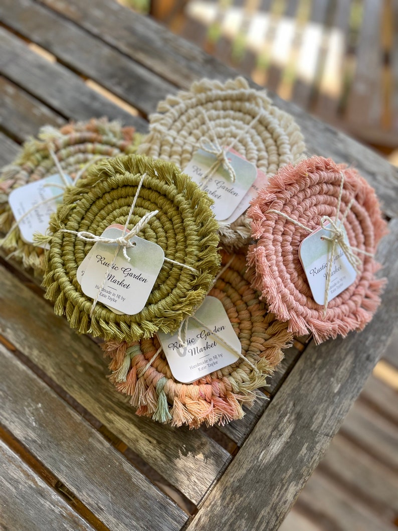 May include: Set of six handmade crocheted coasters with fringe. The coasters are in various colors, including green, pink, and beige. Each coaster has a small square tag with the text "Rustic Garden Market" and "Handmade in MI by Lazie Taylor".