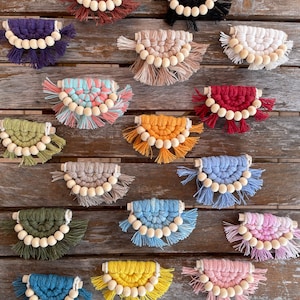 以下が含まれることがあります： 木製のビーズが付いた、カラフルなマクラメの壁掛け18点のセットです。それぞれの壁掛けは、下部にフリンジが付いた半円形をしています。色は、赤、白、青、緑、黄、紫などがあります。