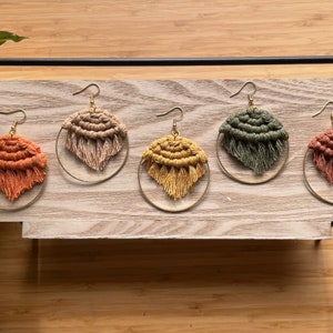 May include: Five pairs of handmade macrame hoop earrings in various colors, including orange, beige, yellow, green, and pink. The earrings are displayed on a wooden surface.