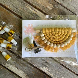 Puede incluir: Un conjunto de pequeñas botellas de vidrio con tapas doradas y goteros blancos, junto con un difusor de aceite para ventilación de automóvil de macramé amarillo y marrón con cuentas de madera. El difusor tiene una etiqueta que dice "CAR VENT OIL DIFFUSER" y "Etsy.com/shop/RusticGardenMarket".