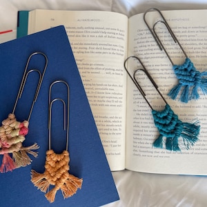 May include: Three handmade macrame bookmarks with colorful tassels attached to silver paper clips. The bookmarks are in shades of pink, orange, and teal.