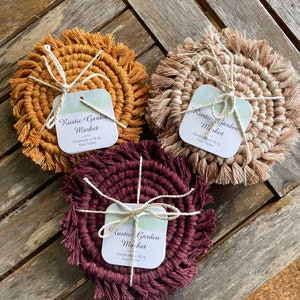 Puede incluir: Tres posavasos de crochet hechos a mano con flecos en burdeos, beige y amarillo mostaza. Cada posavasos tiene una etiqueta cuadrada adjunta con el texto "Rustic Garden Market" y "Handmade in NY by Katie Taylor".