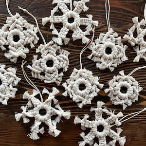 May include: A collection of handmade macrame snowflake ornaments in a natural off-white color. Each snowflake features a circular center and intricate knotting, with fringed edges. They are displayed on a dark wooden surface, ready for hanging.