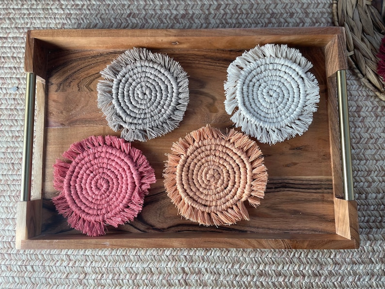 May include: Four round crocheted coasters with fringe. Two are beige, one is peach, and one is pink.