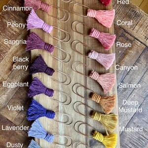 May include: A set of 14 gold metal paper clips with colorful yarn tassels. The tassels are arranged in a gradient from red to yellow, with colors including ruby, cinnamon, peony, sangria, black berry, eggplant, violet, lavender, dusty lavender, deep mustard, mustard, sunflower, and bright yellow.