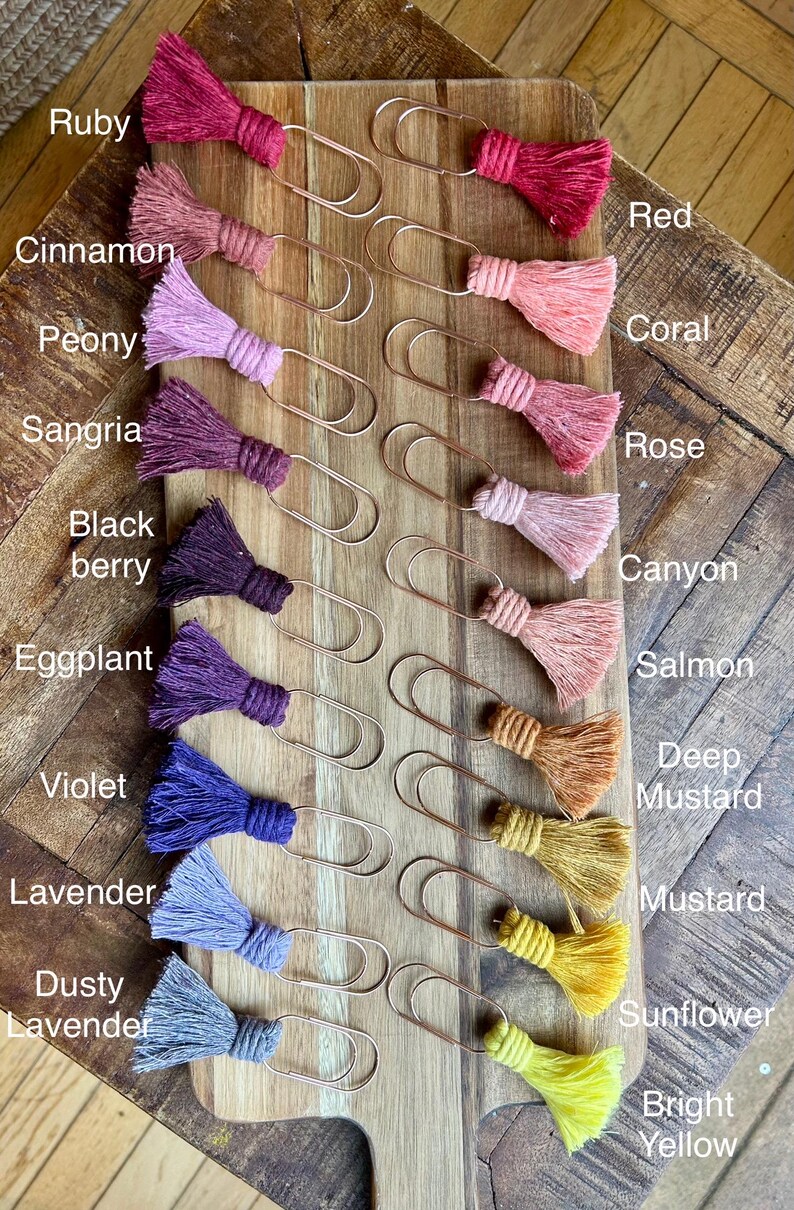 May include: A set of 14 gold metal paper clips with colorful yarn tassels. The tassels are arranged in a gradient from red to yellow, with colors including ruby, cinnamon, peony, sangria, black berry, eggplant, violet, lavender, dusty lavender, deep mustard, mustard, sunflower, and bright yellow.
