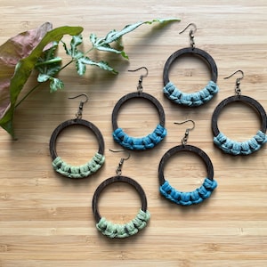 May include: Six pairs of wooden hoop earrings with a woven design. The hoops are brown and the woven design is in shades of green and blue.