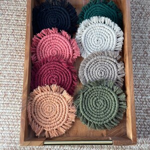 May include: A set of eight round crocheted coasters in a wooden tray. The coasters are in various colors, including black, green, cream, pink, red, and peach.