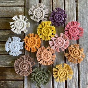 Op de afbeelding: Een verzameling van 12 handgemaakte macramé-onderzetters in verschillende kleuren, waaronder wit, beige, oranje, roze, paars, groen en geel. De onderzetters zijn in een cirkel op een houten oppervlak gerangschikt.