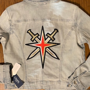 May include: Light blue denim jacket with a large patch featuring a gold, red, and black starburst design with crossed swords. The jacket has a button closure and a tag from the brand Chaps.