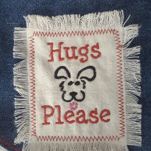 May include: A denim patch with a white, fringed square embroidered with the words "Hugs Please" in red. A black and pink dog face is embroidered between the words. The patch is sewn onto a blue denim background.