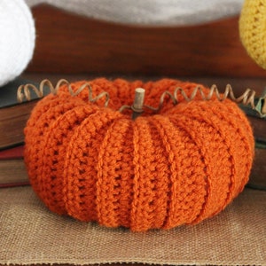 May include: A crocheted orange pumpkin with a wooden stem, sitting on a burlap surface with books and pine cones.