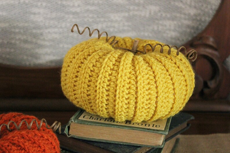 May include: A crocheted yellow pumpkin with a brown stem sits on top of a stack of books. The top book has the text "Charlotte Bronte" printed on the spine.
