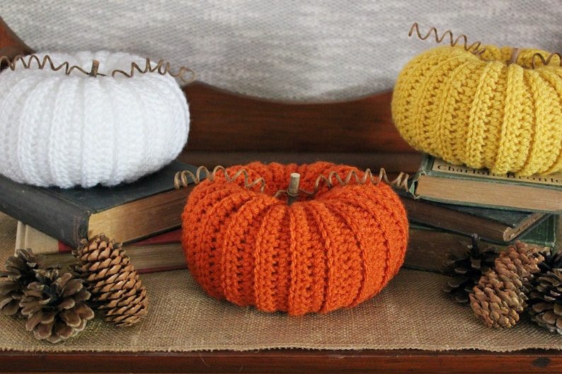 May include: Three crocheted pumpkins in white, orange, and yellow, sitting on a stack of old books with pine cones on a burlap surface.