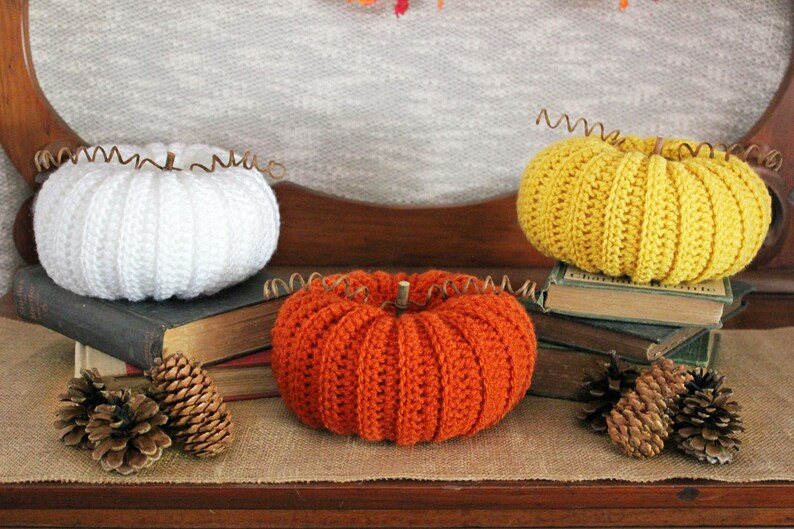 May include: Three crocheted pumpkins, one white, one orange, and one yellow, are displayed on a wooden surface. Each pumpkin has a wire stem and is sitting on a stack of old books. There are pine cones on the surface in front of the pumpkins.
