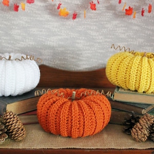 May include: Three crocheted pumpkins in white, orange, and yellow, with brown wire stems. The pumpkins are sitting on a stack of old books and a burlap surface.