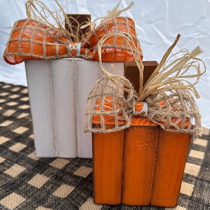 May include: Two wooden pumpkin decorations. One is painted white, the other orange. Both have a burlap bow with orange mesh and raffia accents. A small ribbon with pumpkins is attached to the bow. The pumpkins are on a black and white checkered surface.