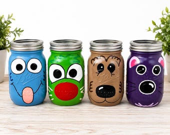 May include: Four glass mason jars with silver metal lids, each painted with a different character face. The blue jar has a tongue out, the green jar has a red nose, the tan jar features a dog-like face, and the purple jar has pink ears.