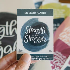 May include: A deck of memory cards titled Strength in the Struggle, featuring a dark blue painted circular brushstroke design on a white background. The text includes Memory Cards, Strength in the Struggle, Lauren Ibach, ESV, and Verses from the Study.
