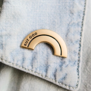 May include: A gold-toned metal pin shaped like a semi-circular rainbow arch. The surface features a smooth, polished finish with a thin engraved line following the curve. The text "BUT GOD" is clearly stamped in black capital letters on the left side.