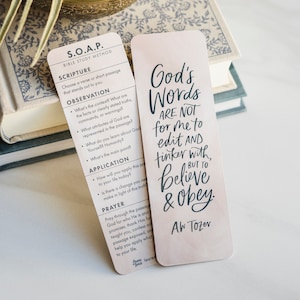 May include: A pink and white bookmark with a quote about God's words. The quote reads "God's Words Are Not For Me To Edit And Tinker With, But To Believe & Obey." The quote is attributed to A.W. Tozer.