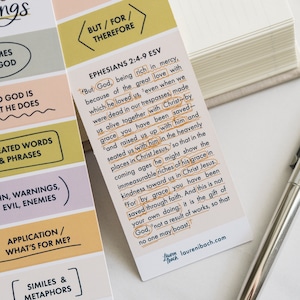 May include: A bookmark with a yellow, orange, pink, and purple striped background. The bookmark features a Bible verse from Ephesians 2:4-9 in the ESV translation. The text is written in black with a yellow outline. The bookmark also includes the words "But/For/Therefore" in a yellow box at the top. The bookmark is designed by Lauren Thach and includes her website address, laurenibach.com.