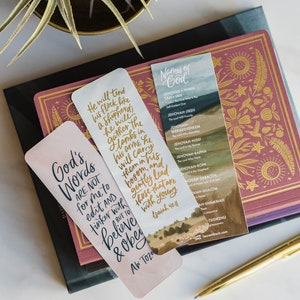 May include: Two bookmarks with inspirational quotes and a list of names of God. The first bookmark is pink with gold lettering and the quote "God's Words are not for me to edit and tinker with but to believe & obey." The second bookmark is blue with gold lettering and the names of God in Hebrew and English.
