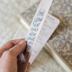 May include: A white bookmark with blue text that reads "God's Words are not for me to edit and tinker with, but to live and obey."  The text is written in a cursive font.  The bookmark also includes the text "A.W. Tozer" at the bottom.