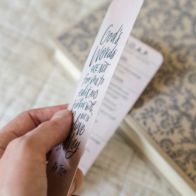 May include: A white bookmark with blue text that reads "God's Words are not for me to edit and tinker with, but to live and obey."  The text is written in a cursive font.  The bookmark also includes the text "A.W. Tozer" at the bottom.