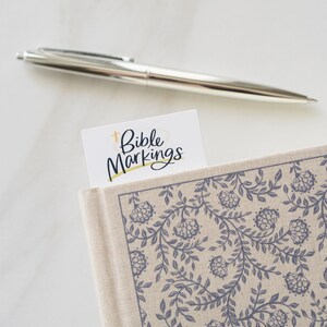 May include: A white bookmark with the text "Bible Markings" in black lettering and a gold cross symbol.