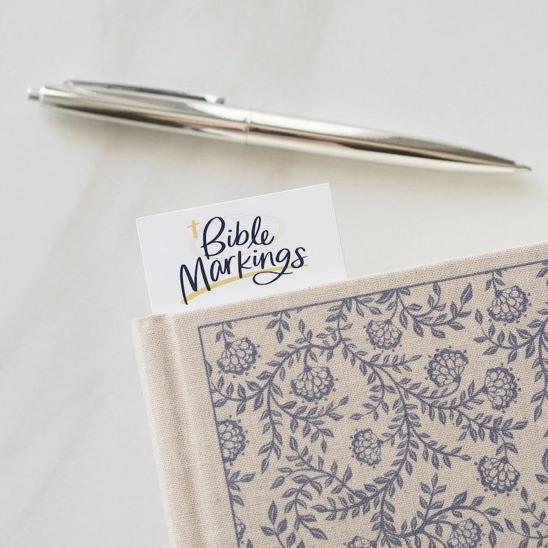 May include: A white bookmark with the text "Bible Markings" in black lettering and a gold cross symbol.