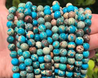 Puede incluir: Una hebra de cuentas de jaspe redondas de color azul turquesa, marrón y blanco. Las cuentas tienen un patrón moteado.