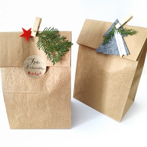 Könnte beinhalten: Zwei braune Papiertüten mit Schneeflockenmustern. Eine Tüte hat einen roten Stern und ein Etikett mit der Aufschrift "Frohe Weihnachten". Die andere Tüte hat einen grauen Filz-Weihnachtsbaum mit einem Etikett mit der Aufschrift "Frohe Weihnachten".