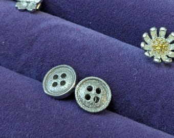May include: Two silver metal buttons with four holes each. The buttons are on a purple velvet surface.