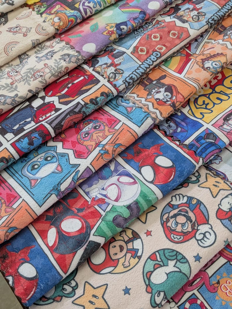 May include: A stack of colorful fabric featuring comic book panels and cartoon characters. Designs include Spider-Man, Mario, Lightning McQueen, and other popular characters. The fabric has a soft, plush texture and a variety of bright colors.