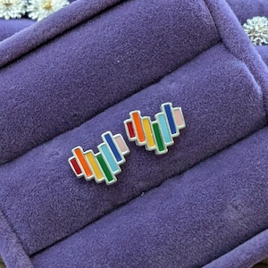 May include: A pair of rainbow heart-shaped earrings on a purple velvet background. The earrings are made of silver and have a rainbow enamel finish.