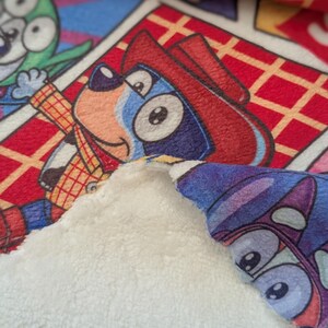 May include: A close-up of a colorful blanket featuring cartoon characters. The blanket has a soft, white, plush backing. The front displays a comic-style pattern with characters in red, blue, green, and yellow.