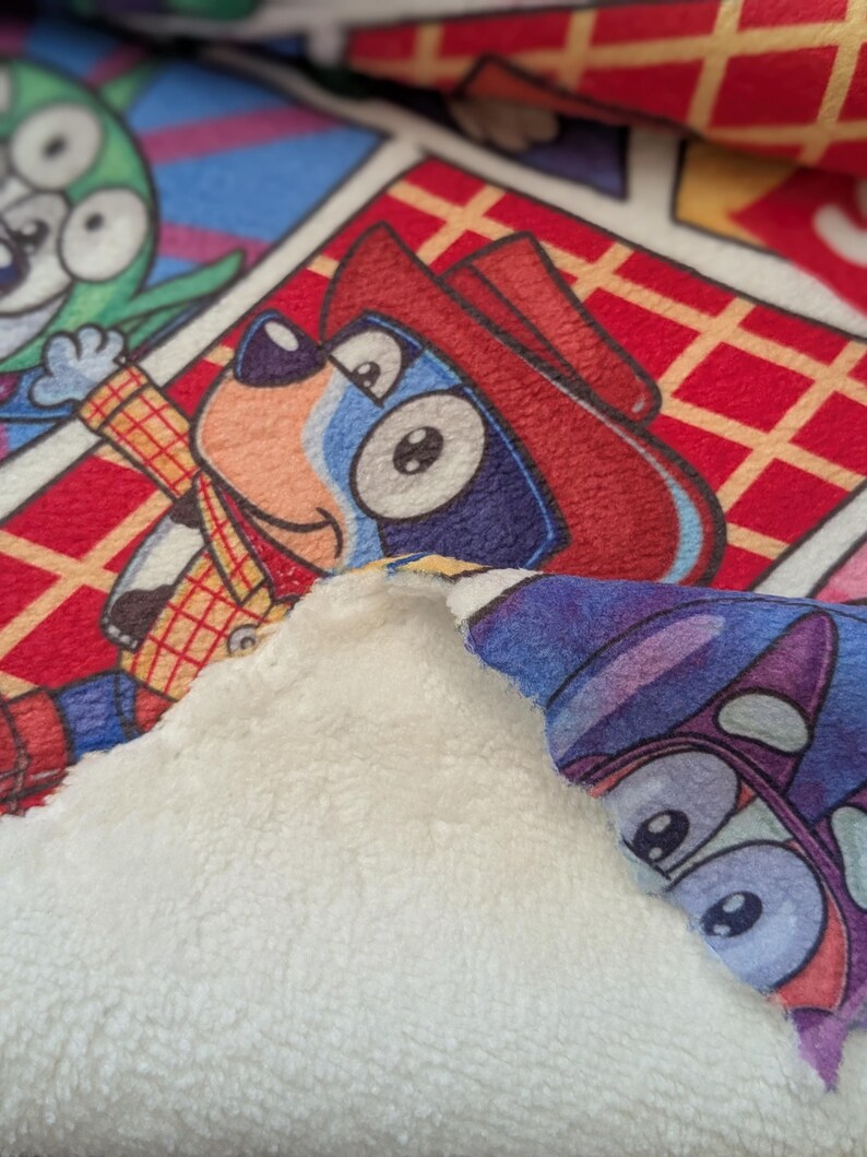 May include: A close-up of a colorful blanket featuring cartoon characters. The blanket has a soft, white, plush backing. The front displays a comic-style pattern with characters in red, blue, green, and yellow.
