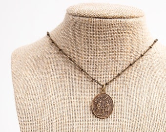 May include: A brown necklace with a round pendant featuring a detailed design on a burlap jewelry display stand.