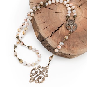 May include: A white and brown agate rosary with a bronze crucifix and a decorative centerpiece. The rosary is displayed on a wooden slice.
